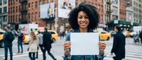 Young African American woman with curly hair smiling and holding blank white sign on busy urban street with yellow taxis and city crowd.