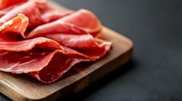 Thinly sliced cured prosciutto ham arranged on wooden cutting board against dark background, macro food photography showing meat texture and marbling.