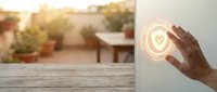 Hand holding glowing heart symbol with warm light on blurred outdoor terrace background for wellness and care concepts.