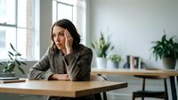 Young professional woman sitting at wooden desk in modern office space with plants, looking thoughtful and contemplative during work break.