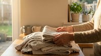 Woman in cozy sweater folding clean laundry clothes on wooden table near sunny window with plants creating warm domestic atmosphere for home organization.