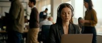 Young woman with long brown hair and light skin tone wearing headphones and dark blazer, working on laptop in modern office environment with blurred colleagues in background, representing remote work, concentration, and professional communication.