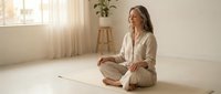 Young Caucasian woman in beige outfit practicing mindfulness meditation sitting cross-legged on yoga mat in bright minimalist living room with natural sunlight.
