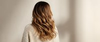 Young woman with long wavy blonde hair wearing beige ribbed sweater from behind against neutral background for fashion and lifestyle concepts.