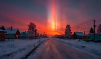 Dramatic winter sunrise over snowy village road with vibrant orange and pink sky creating stunning light pillar phenomenon in rural residential area.