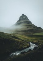 Dramatic mountain peak shrouded in mist with winding river stream below, moody atmospheric landscape featuring green slopes and foggy conditions in wilderness.
