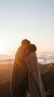 Romantic couple embracing at mountain summit during golden hour sunrise with dramatic clouds and warm sunlight creating intimate silhouette moment.