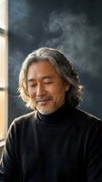 Mature Asian man with gray hair and beard smiling peacefully with eyes closed in black turtleneck sweater by window with natural light and dark background.