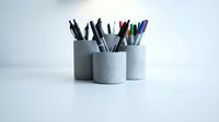 Gray concrete cylindrical pen holders with colorful pens and pencils on white desk surface for modern office workspace organization and stationery storage.