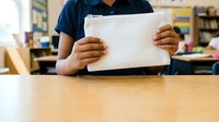 Young student of African descent wearing navy blue shirt holding tablet device at wooden desk in classroom with blurred educational background and bookshelves.