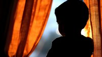 Silhouette of child looking through window at sunset with orange curtains creating dramatic lighting and peaceful contemplative mood. Suitable for childhood themes.