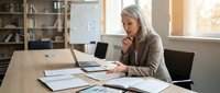 Mature woman with long white hair and light complexion, wearing a beige cardigan, sitting at a wooden desk in a bright office environment, working on a laptop with notebooks and documents spread around her, appearing focused and thoughtful while working on business tasks.