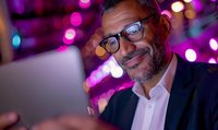Middle-aged Hispanic businessman with glasses smiling while looking at digital tablet screen in evening setting with vibrant purple and blue bokeh lights background.