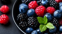 Mixed berries including raspberries, blueberries, and blackberries arranged in dark bowl with mint leaf garnish on slate surface.