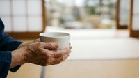 Close-up of mature man's hands holding white ceramic mug indoors near window with natural light, suitable for beverage, lifestyle and wellness marketing.