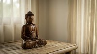 Bronze Buddha statue in meditation pose on wooden table near window with soft natural light creating peaceful zen atmosphere for mindfulness and spiritual practice.