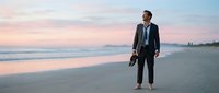 Caucasian male professional in business suit standing barefoot on sandy beach at sunset, holding shoes, contemplating ocean horizon during golden hour.