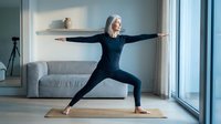 Mature woman with long gray hair, Caucasian ethnicity, performing warrior pose yoga exercise on mat in bright modern living room with natural light from windows and contemporary furniture.