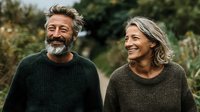 Mature couple laughing together outdoors, wearing cozy sweaters against natural garden background, conveying joy and connection in retirement.