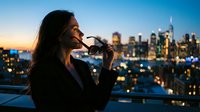 Woman with long dark hair and olive complexion, wearing business attire, adjusting eyeglasses while overlooking illuminated city skyline at dusk. Urban professional lifestyle and evening cityscape.