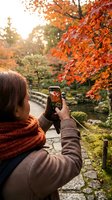 Woman with dark hair photographed from behind wearing brown sweater and orange scarf, holding smartphone to capture vibrant autumn foliage in garden setting with stone pathway and water feature.
