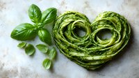 Heart shaped green pasta with pesto sauce and fresh basil leaves on light textured background. Creative food styling for healthy Italian cuisine.