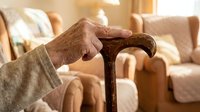 Elderly person gripping a wooden walking cane indoors, soft living room background, concept of aging, mobility aid, and senior care.