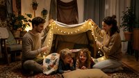 Happy family building cozy blanket fort with fairy lights in warm living room. Parents and children creating magical indoor camping experience together.
