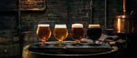 Craft beer flight featuring four glasses with varying amber and dark brews displayed on wooden barrel in rustic brewery setting with brick walls and copper equipment for tasting and hospitality.