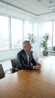 Mature male executive in dark blazer seated at wooden conference table in modern high-rise office with floor-to-ceiling windows overlooking cityscape, potted plants, and minimalist decor.