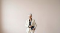 Senior woman with gray hair, wearing cream blazer and dark top, holding tablet device against neutral beige background for corporate and business use.