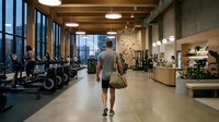 Modern fitness center interior with athletic man walking through spacious gym facility featuring exercise equipment, wooden beams, and urban windows.
