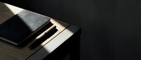Closed black notebook and beige folder stacked on wooden desk with dramatic sunlight casting sharp shadows, suitable for business and office design projects.
