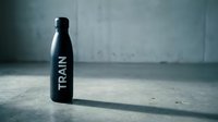 Black water bottle with train text on concrete floor in industrial setting with dramatic lighting.