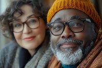 Happy mature African American man with gray beard and glasses wearing orange knit hat smiling with Caucasian woman partner in cozy indoor setting.