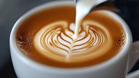 Barista pouring steamed milk into coffee cup creating leaf pattern latte art, close-up view showing intricate design and rich brown crema surface.
