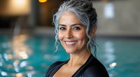 Confident mature woman with silver gray hair smiling at indoor pool, wearing black athletic wear. Natural aging and wellness lifestyle concept for active seniors.