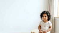 Young child with curly dark hair and medium skin tone wearing white t-shirt, sitting indoors by window with natural light, suitable for family, parenting, childhood, and lifestyle editorial content.