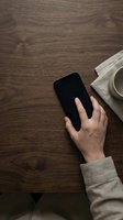 Hand holding black smartphone on wooden desk with coffee cup and notebook, overhead workspace view for business and technology concepts.