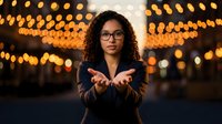 Young African American businesswoman with curly hair and glasses extending hands forward against warm bokeh lights background at night.
