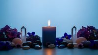 Dark blue candle burning with flame surrounded by colorful gemstones, crystals and decorative stones on blue background for meditation and wellness.