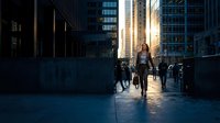 Young professional woman walking confidently through urban business district during golden hour with modern skyscrapers and city commuters in background.