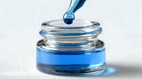 Blue liquid droplet falling from dropper into small glass container with vibrant blue solution against white background. Perfect for cosmetics or laboratory concepts.