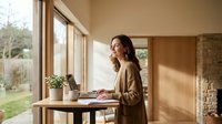 Young professional woman working on laptop at wooden desk by large windows in modern home office with natural lighting and plants.