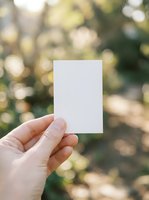 Hand holding a blank white card outdoors with a blurred green bokeh background, copy space for text or branding mockup.