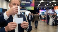 Middle-aged Caucasian male professional in business suit holding blank ID badge at conference or trade show event with attendees and exhibition booths in background.