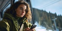 Young woman with curly hair using smartphone while traveling by train through snowy mountain landscape. Winter journey and mobile communication concept.