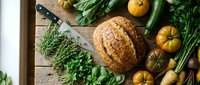 Fresh organic vegetables and artisan bread on rustic wooden cutting board with herbs and knife for healthy cooking and meal preparation.