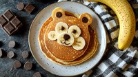 Cute bear-shaped pancake decorated with banana slices and chocolate chips, served on gray plate. Creative breakfast for kids with fresh banana and chocolate pieces.