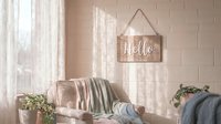 Cozy pastel living room with a rustic wooden Hello sign hanging on a brick wall, soft natural light, and lush green plants.